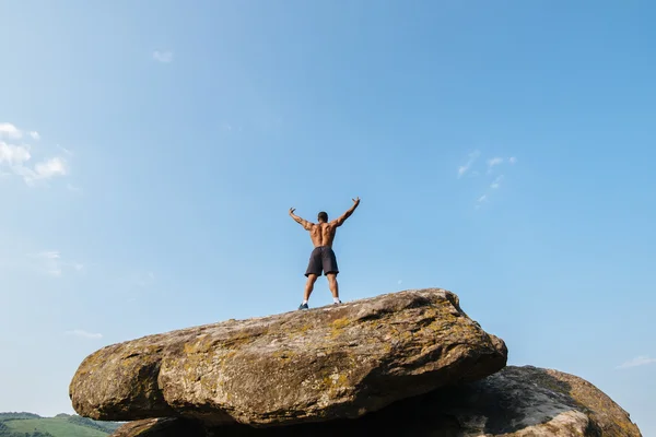 Achteraanzicht van sterke zwarte bodybuilder poseren op de rots tegen blauwe bewolkte hemel — Stockfoto