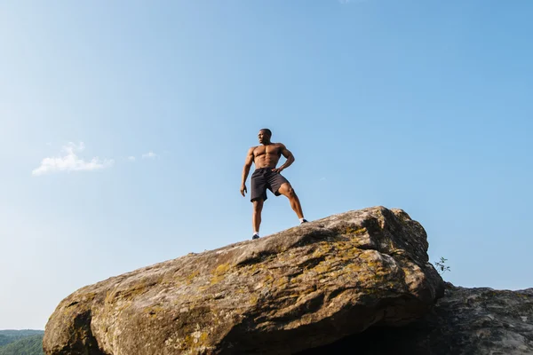 岩の上にポーズ裸の胴体を持つ強い黒人アフリカ系アメリカ人男性ボディービルダーの肖像画。青い曇り空の背景 — ストック写真