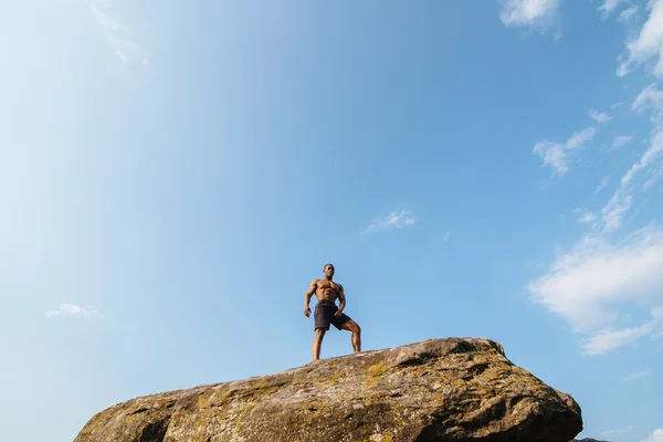 Svart afrikansk amerikansk man bodybuilder poserar på klippan med blå himmel bakgrund — Stockfoto