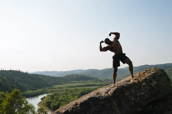 岩の上にポーズ裸の胴体を持つアフリカ系アメリカ人ボディービルダーのシルエット.緑の山の自然の背景 — ストック写真