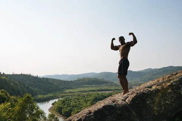 Retrato al aire libre del culturista afroamericano con torso desnudo posando sobre la roca. Verde montaña naturaleza fondo —  Fotos de Stock