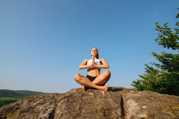 Portret van sexy jonge atletische vrouw met gespierde lichaam beoefenen van yoga op de rots. Blauwe hemel achtergrond — Stockfoto