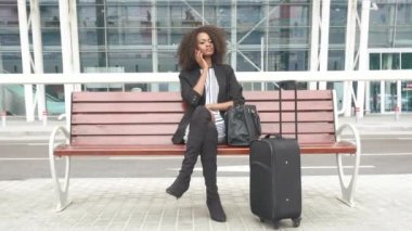 Havaalanında bankta oturmuş ve telefonda konuşurken muhteşem genç gülümseyen Afro-Amerikan iş kadını. Kentsel cam duvar arka plan