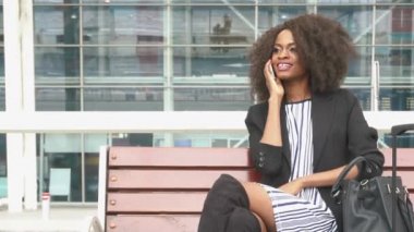 Havaalanında bankta oturmuş ve bir uçuş için beklerken telefonda konuşurken muhteşem gülümseyen Afro-Amerikan iş kadını. Kentsel cam duvar arka plan