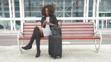 Bir uçuş için beklerken, Havaalanı, smartphone cep telefonu ile mesaj bankta oturmuş muhteşem gülümseyen Afro-Amerikan iş kadını.