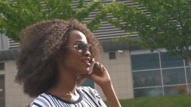 Güneş gözlüklü, cep telefonuyla konuşan ve gülümseyen genç Afro-Amerikan iş kadını.
