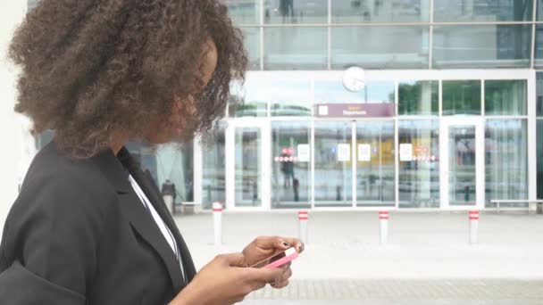 Boční pohled atraktivní černé podnikání lady SMS na telefonu při čekání na letadlo na letišti. Městské skleněná stěna na pozadí — Stock video