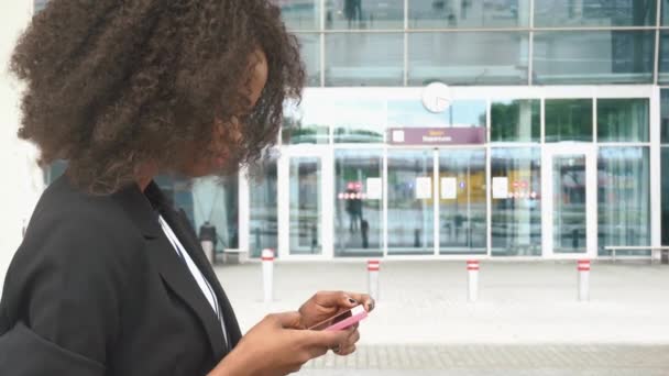 Vue latérale de la belle femme d'affaires afro-américaine textant au téléphone en attendant un vol à l'aéroport. Mur de verre urbain sur fond — Video