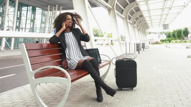 Portret van aantrekkelijke jonge lachende Afro-Amerikaanse zakenvrouw zittend op het bankje op de luchthaven en praten op mobiele telefoon — Stockvideo