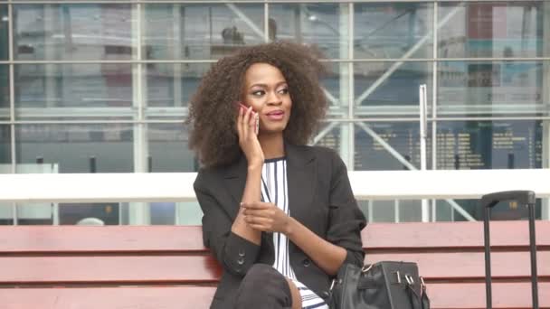 Gros plan d'une jolie jeune femme d'affaires afro-américaine souriante assise sur le banc de l'aéroport et parlant sur son téléphone portable. Fond mural en verre urbain — Video