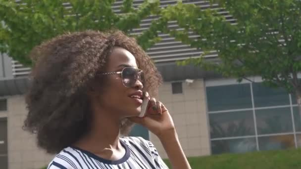 Joven atractiva mujer de negocios afroamericana en gafas de sol hablando por teléfono móvil y sonriendo — Vídeos de Stock