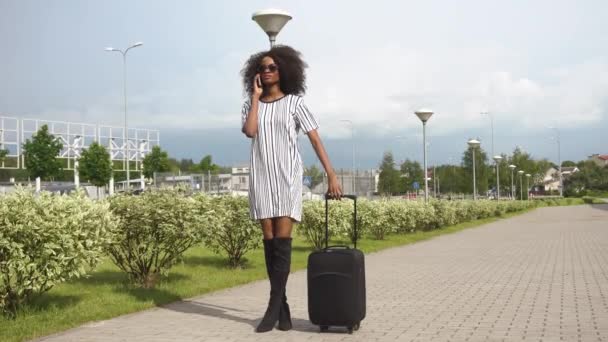Fullängds utsikt över eleganta leende afroamerikanska affärskvinna med solglasögon på flygplatsen med hennes bagage. Black Lady pratar med telefon i väntan på en flygning. Affärs resekoncept — Stockvideo