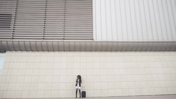 Elegante schlanke Afroamerikanerin mit Gepäck an der massiven Flughafenmauer. weiße strukturierte Wand Hintergrund. Bereiten Sie sich auf die Reise vor. Zerbrechliche Frau und massive moderne Architektur — Stockvideo