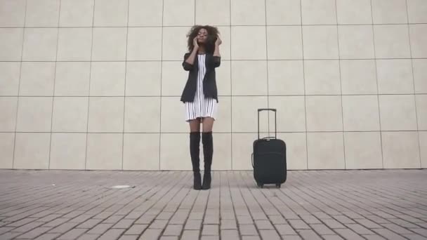 Elegante mujer afroamericana delgada delgada con equipaje hablando por teléfono en el aeropuerto. Fondo de pared con textura blanca. Preparándose para el viaje. Colores monocromáticos — Vídeo de stock