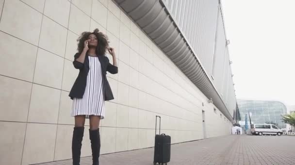 Atractiva mujer de negocios afroamericana con equipaje hablando por teléfono y esperando a subir al avión. Aeropuerto pared blanca fondo — Vídeo de stock