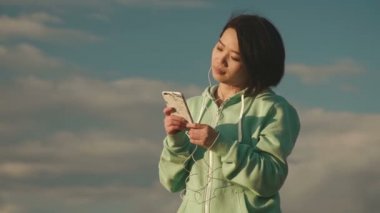 Şehvetli genç Asyalı kadın dinle müzik kulaklık ve dans etmek için. Harika mavi gökyüzü arka plan, ağır çekim