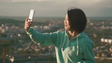 Güzel genç Japon kadın selfie açık park onun telefon ve gösterileri Tamam bulanık şehir arka plan fotoğraf makinesini kullanarak alma