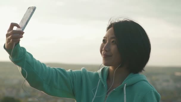 Красивая японская женщина делает селфи на открытом воздухе в парке, используя свой телефон и подмигивает — стоковое видео