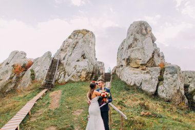 Şık mavi yakışıklı damat takım elbise kucaklayan beyaz giyimli gelin gül buketi artalanları olarak görkemli dağ manzarası ile büyük kayalar holding