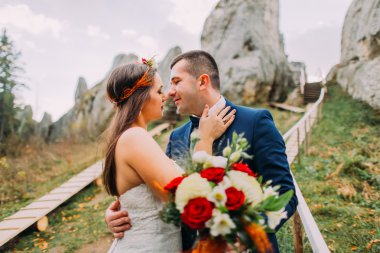 Şık mavi yakışıklı damat öpüşme beyaz giyimli gelin gül buketi artalanları olarak görkemli dağ manzarası ile büyük kayalar üzerinde tutarak uygun görünümü kadar kapatın