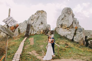 Şık mavi yakışıklı damat takım elbise kucaklayan beyaz giyimli gelin gül buketi büyük kayalar ve artalanları olarak eski mesaj görkemli açık peyzaj üzerinde tutan