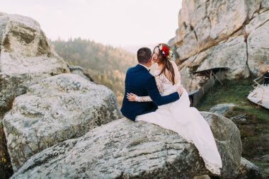 Zarif damat sevgiyle görkemli dağ kayalık manzaraya bakarak şirin baş çelenk ile onun sevimli beyaz giyimli gelin tutan şık mavi elbiseli. Arkadan Görünüm