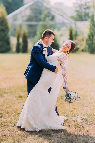 Bräutigam im stilvollen blauen Anzug hält schöne Braut mit weißem Brautkleid draußen auf dem Rasen. Gewächshaus im Hintergrund — Stockfoto