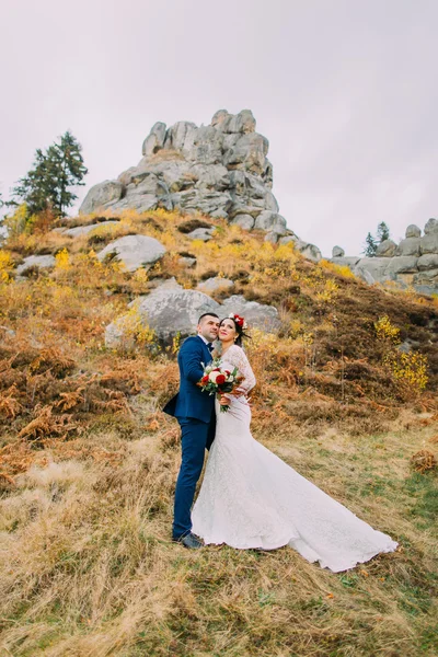 Guapo novio en elegante traje azul abrazando novia vestida blanca sosteniendo ramo de rosas en majestuoso paisaje al aire libre con grandes rocas como fondo —  Fotos de Stock