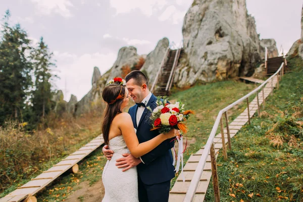 Knappe bruidegom in stijlvolle blauwe pak zoenen zijn wit gekleed bruid bedrijf boeket rozen op de majestueuze berglandschap met enorme rotsen als backround — Stockfoto