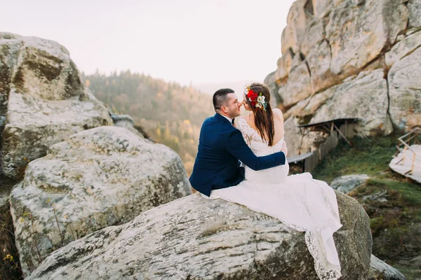 在时尚蓝色优雅新郎适合心疼地抱着可爱的头花圈的白色穿着的新娘上雄伟的山脉作为背景的岩石景观。后视图 — 图库照片