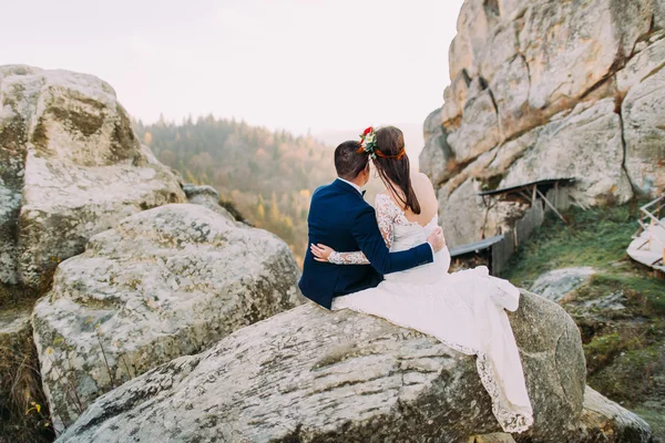 Elegante bruidegom in stijlvolle blauwe pak liefdevol holding wit gekleed bruid met schattige hoofd krans op zoek op majestueuze berg rotsachtige landschap. Achteraanzicht — Stockfoto