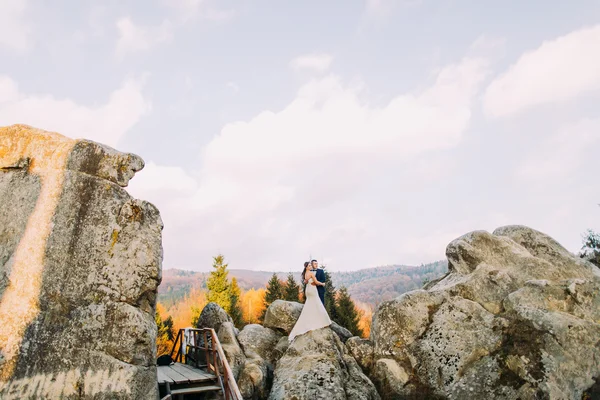 Beau marié en costume bleu élégant embrassant mariée habillée blanche sur paysage extérieur idyllique avec de gros rochers et ciel nuageux comme backround — Photo