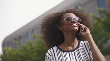 Sevimli genç Afro-Amerikan iş kadın telefonla konuşuyor ve gülümseyen güneş gözlüğü. Cityscape arka plan