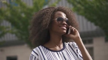 Kendine güvenen genç Afro-Amerikan iş kadını telefonla konuşurken ve gülümseyen güneş gözlüğü. Cityscape arka plan