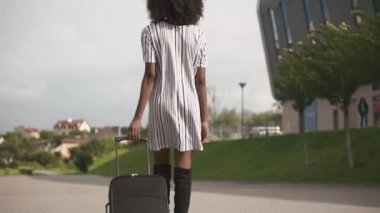 Arkadan görünüşü zarif Afro-Amerikan iş kadını şık kısa elbise onun Bagaj ile havaalanına gidiyor. İş seyahat kavramı