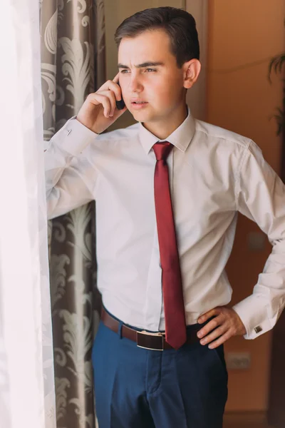 Pengusaha muda bermasalah menggunakan ponsel yang berdiri di dekat jendela kamar hotel — Stok Foto