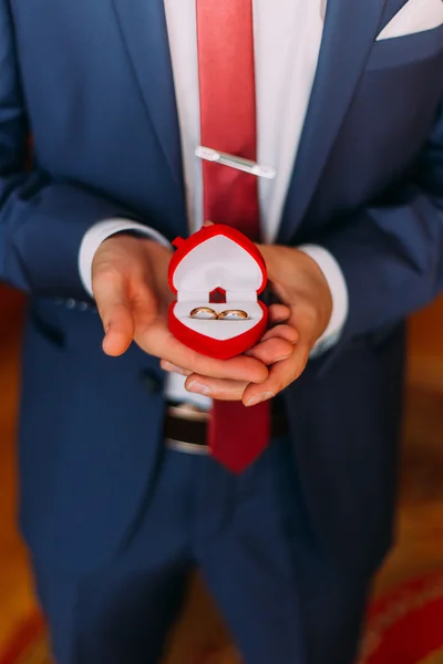 Bonito homem vestido de luxo em azul elegante segurando caixa em forma de coração vermelho com anéis de casamento. Close-up — Fotografia de Stock