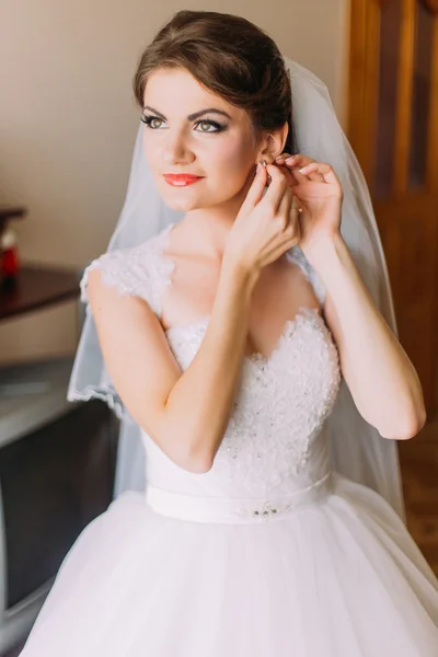 Bela noiva em vestido de noiva branco coloca no brinco. Retrato feminino em vestido de noiva para o casamento. Bonita senhora dentro de casa — Fotografia de Stock