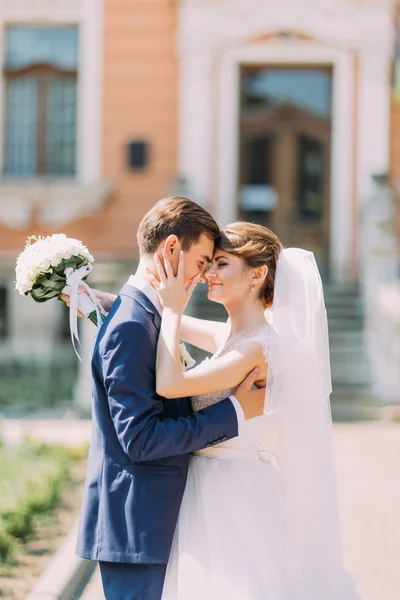 Frisch verheiratetes Paar posiert im sonnigen Park an der Gasse. Bräutigam und Braut halten sich sanft. Altbau als Hintergrund — Stockfoto