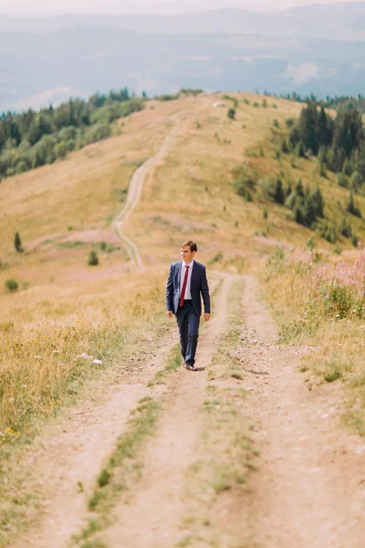 Jovem de terno elegante andando na trilha por campo de verão com colinas no fundo — Fotografia de Stock