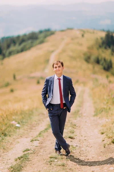 Jeune homme en costume élégant debout sur la piste à travers le champ d'été avec des collines à l'arrière-plan — Photo