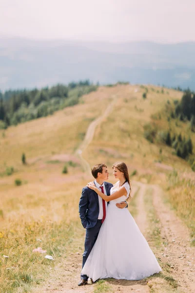 Pareja romántica posando en el sendero a través del campo soleado amarillo con colinas forestales como fondo —  Fotos de Stock