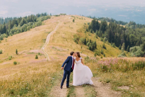 Romantisches Paar küsst sich auf dem Weg über gelbes Sonnenfeld mit Waldhügeln als Hintergrund — Stockfoto