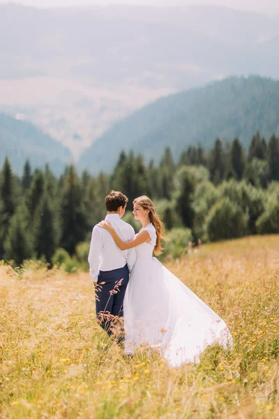 Vackra unga bröllopsparet går på soliga fält med magnifika skogen kullar i bakgrunden. Bakifrån — Stockfoto