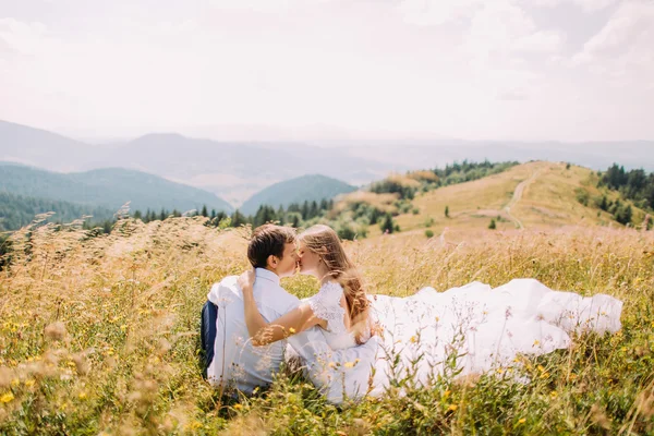 Romántica pareja joven sentada en la hierba en el campo soleado amarillo con colinas forestales distantes como fondo —  Fotos de Stock