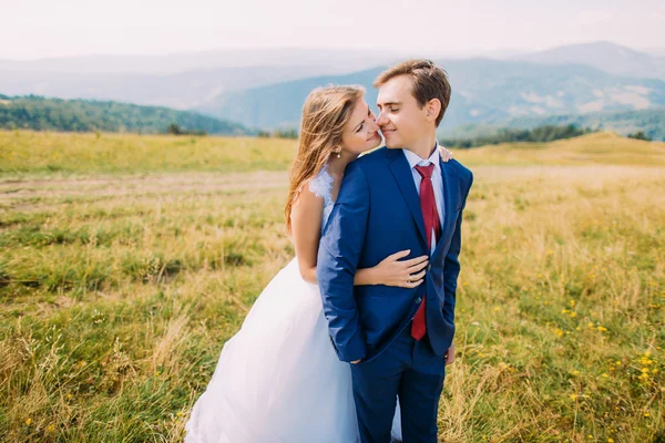 Jeune couple de mariage posant sur un champ ensoleillé avec des collines forestières lointaines comme arrière-plan. Mariée ludique tenant son marié — Photo