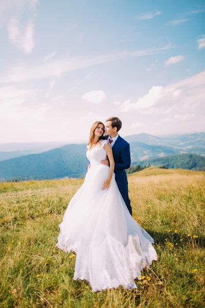 Νέοι ρομαντικό γάμο ζευγάρι ποζάρει στο πεδίο ηλιόλουστη γρασίδι με μακρινό Δασικοί λόφοι και ουρανό ως φόντο — Φωτογραφία Αρχείου