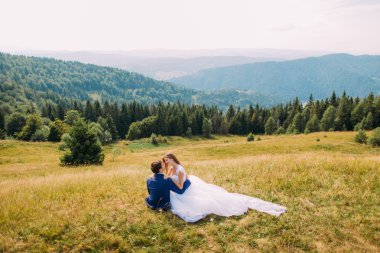 Romantik Çift arka plan olarak görkemli forest hills ile güneşli alan sarı çimlere oturup. Arkadan Görünüm