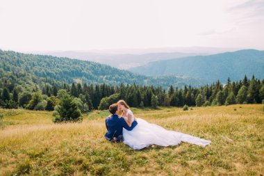Arka plan olarak görkemli orman tepeleri ile güneşli alanda sarı çim üzerinde oturan onu sevgi dolu damat elinde Romantik gelin