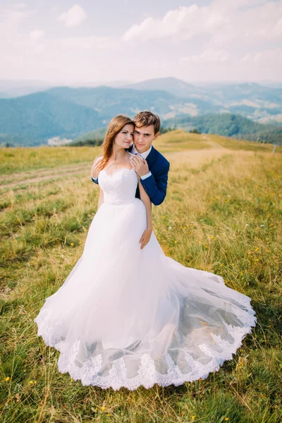 Νέοι ρομαντικό γάμο ζευγάρι ποζάρει στο πεδίο ηλιόλουστη γρασίδι με μακρινό Δασικοί λόφοι και ουρανό ως φόντο — Φωτογραφία Αρχείου